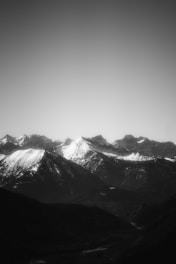 A black and white image of rugged mountain peaks with snow-capped summits under a clear sky, creating a dramatic contrast between the bright snow and dark rock faces.
