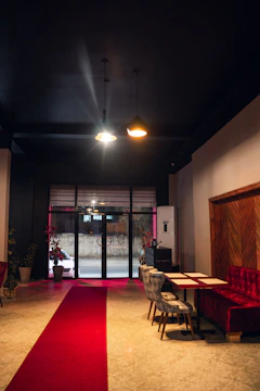 A dimly lit interior space with a red carpet leading to large glass doors. The area features red velvet seating on the right and two gray chairs at a table. Two pendant lights hang from the dark ceiling, casting soft illumination. Comfortable and stylish decor elements include potted plants near the entrance and a wooden accent wall.