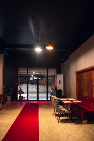 A dimly lit interior space with a red carpet leading to large glass doors. The area features red velvet seating on the right and two gray chairs at a table. Two pendant lights hang from the dark ceiling, casting soft illumination. Comfortable and stylish decor elements include potted plants near the entrance and a wooden accent wall.