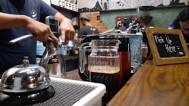 A barista uses a manual espresso machine to prepare coffee on a countertop. In the foreground, a glass pitcher filled with freshly brewed coffee sits beside a small bell. The background features a chalkboard sign that reads 'Pick Up Here', and various coffee-making equipment, including a grinder and a carton of almond milk. The setting appears to be a cozy, rustic-style coffee shop.