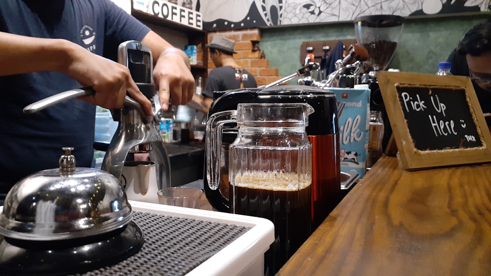 A barista uses a manual espresso machine to prepare coffee on a countertop. In the foreground, a glass pitcher filled with freshly brewed coffee sits beside a small bell. The background features a chalkboard sign that reads 'Pick Up Here', and various coffee-making equipment, including a grinder and a carton of almond milk. The setting appears to be a cozy, rustic-style coffee shop.