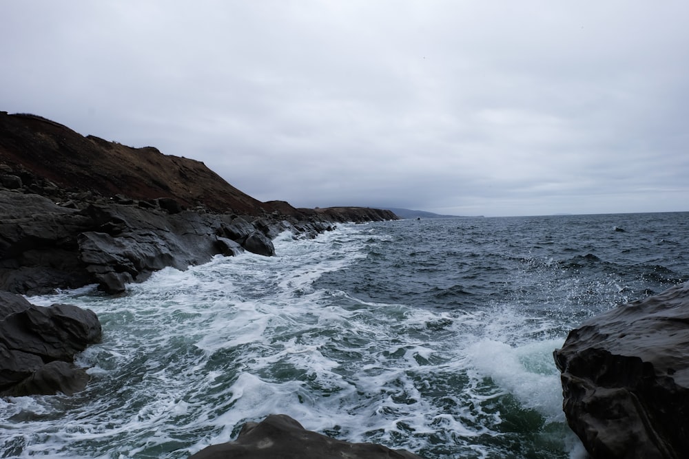 ocean waves crashing on brown rocky mountain under white cloudy sky during daytime