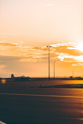 A tranquil scene at an airport during sunset with warm golden hues dominating the sky. The silhouette of an airplane is visible on the tarmac, and street lamps line the pavement. The atmosphere is calm and almost deserted, with a soft glow from the setting sun.