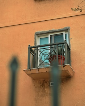 A small balcony with intricate metal railing design is attached to an orange textured building wall. A potted plant with long leaves sits on the balcony beside a window with closed blinds.