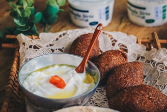A silver bowl filled with creamy white yogurt topped with a small amount of orange garnish and olive oil, accompanied by a small brown spoon. The scene includes several pieces of dark, crispy-fried food items, likely falafel, resting on a lace-lined tray. In the background, there are jars of what appears to be yogurt or labneh and two stalks of green leaves for decoration.