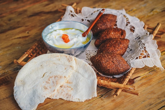 A rustic wooden table holds a woven tray topped with a piece of lace fabric. On the tray, there are three dark, crispy pastries, possibly kibbeh, positioned next to a bowl of creamy yogurt garnished with a splash of olive oil and a small red pepper piece. Beside the bowl, there is a piece of flatbread with a portion torn off.