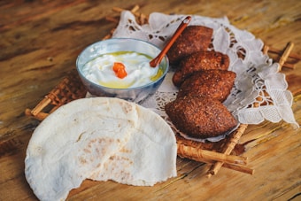A rustic wooden table holds a woven tray topped with a piece of lace fabric. On the tray, there are three dark, crispy pastries, possibly kibbeh, positioned next to a bowl of creamy yogurt garnished with a splash of olive oil and a small red pepper piece. Beside the bowl, there is a piece of flatbread with a portion torn off.