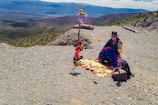A person wearing traditional clothing sits on a rocky mountain path with colorful handmade crafts and textiles laid out on a mat. Brightly colored decorations are hung on a rustic frame behind them, and expansive mountainous landscape stretches into the distance.