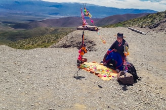 A person wearing traditional clothing sits on a rocky mountain path with colorful handmade crafts and textiles laid out on a mat. Brightly colored decorations are hung on a rustic frame behind them, and expansive mountainous landscape stretches into the distance.
