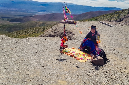 A person wearing traditional clothing sits on a rocky mountain path with colorful handmade crafts and textiles laid out on a mat. Brightly colored decorations are hung on a rustic frame behind them, and expansive mountainous landscape stretches into the distance.