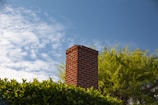 A red brick chimney extends above lush green foliage, under a bright blue sky with scattered white clouds.