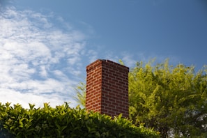 A red brick chimney extends above lush green foliage, under a bright blue sky with scattered white clouds.