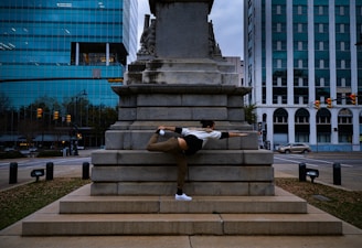 A person performs a flexible balance pose against a large stone monument in an urban environment. The scene includes surrounding modern office buildings with glass windows and some traffic signals.