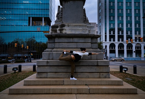A person performs a flexible balance pose against a large stone monument in an urban environment. The scene includes surrounding modern office buildings with glass windows and some traffic signals.