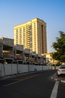 A tall, beige-colored residential or office building stands prominently in the background, with a partially constructed building in the foreground surrounded by metal fencing. The construction appears incomplete, with exposed concrete and metal structures. A road curves around the construction site, and a person is walking along the sidewalk beside the fence. There is a car parked on the side of the road, and a tree's foliage is visible on the right side. The sky is clear and blue, suggesting a sunny day.