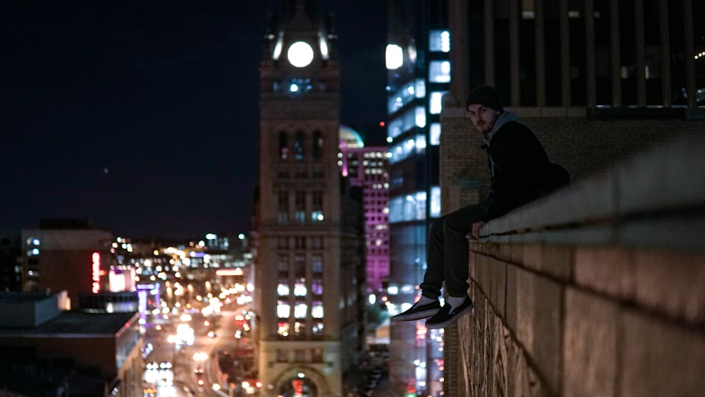 man in black hoodie and black pants sitting on brown wooden fence during night time