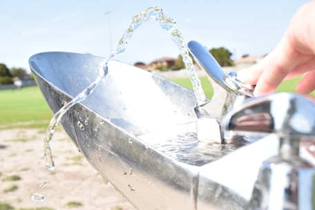 A close-up of a hand operating a silver drinking fountain with water arcing gracefully in mid-air. The background features a park with green grass and trees under a clear blue sky.