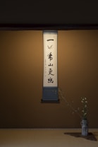 A calligraphy scroll is elegantly displayed on a simple wall, featuring Japanese characters with a minimal decorative design at the top. Below the scroll, a delicate flower arrangement in a rustic vase is placed on a wooden mat, adding a touch of natural beauty to the scene.