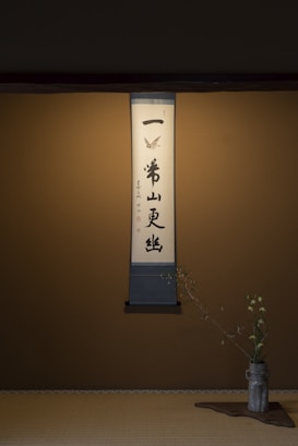 A calligraphy scroll is elegantly displayed on a simple wall, featuring Japanese characters with a minimal decorative design at the top. Below the scroll, a delicate flower arrangement in a rustic vase is placed on a wooden mat, adding a touch of natural beauty to the scene.