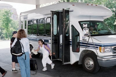 A small bus is parked with its door open, allowing a child with a backpack to step inside. Another child and an adult holding a briefcase are standing nearby. The driver is visible through the window. The setting appears to be a bus stop with trees and a brick building in the background.