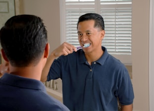 A smiling person brushing their teeth with natural light streaming through a bathroom window.