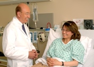A doctor in a white coat stands next to a woman lying in a hospital bed, engaging in a conversation. The patient is smiling and wearing a hospital gown, while medical equipment is visible in the background.