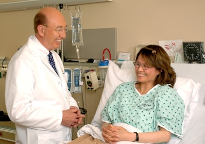 A doctor in a white coat stands next to a woman lying in a hospital bed, engaging in a conversation. The patient is smiling and wearing a hospital gown, while medical equipment is visible in the background.