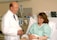 A doctor in a white coat stands next to a woman lying in a hospital bed, engaging in a conversation. The patient is smiling and wearing a hospital gown, while medical equipment is visible in the background.