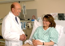 A doctor in a white coat stands next to a woman lying in a hospital bed, engaging in a conversation. The patient is smiling and wearing a hospital gown, while medical equipment is visible in the background.