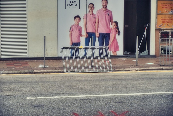 Billboard of a family wearing coordinated pink outfits, displayed on a storefront. The family consists of two adults and two children. In front of the billboard is a metal barrier on the sidewalk. The scene includes an urban street setting with visible signage.