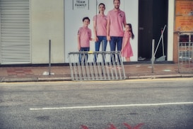 Billboard of a family wearing coordinated pink outfits, displayed on a storefront. The family consists of two adults and two children. In front of the billboard is a metal barrier on the sidewalk. The scene includes an urban street setting with visible signage.