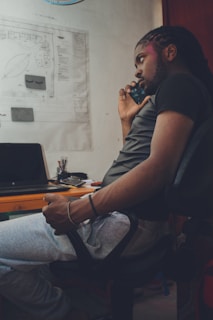 A person with braided hair is sitting in an office chair, holding a smartphone to their ear. They are wearing a black t-shirt and gray sweatpants. On the desk next to them are a laptop, some pens, and papers. A large drawing is pinned to the wall.