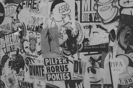 A collage of various posters, stickers, and graffiti covers a surface. The black and white image features diverse designs including cartoon characters, stylized text, and abstract shapes. Some materials appear torn and layered, giving a textured and weathered look. The overall scene conveys a street art aesthetic.