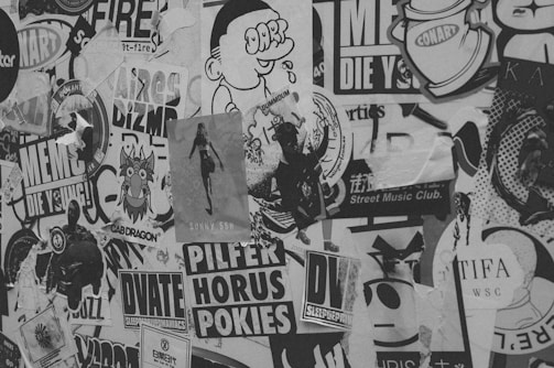 A collage of various posters, stickers, and graffiti covers a surface. The black and white image features diverse designs including cartoon characters, stylized text, and abstract shapes. Some materials appear torn and layered, giving a textured and weathered look. The overall scene conveys a street art aesthetic.