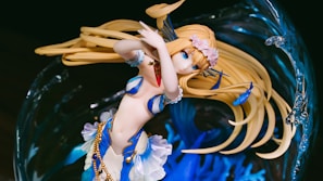 A detailed anime-style figure of a female character with long blonde hair and blue eyes, adorned with floral accessories and golden accents. Her attire is a mix of blue and white with seashell motifs, suggesting an oceanic or aquatic theme. She is surrounded by an abstract background resembling swirling water with intricate details.