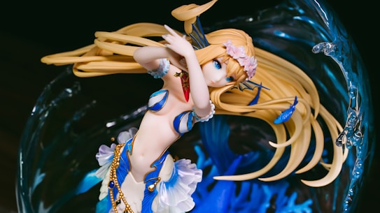 A detailed anime-style figure of a female character with long blonde hair and blue eyes, adorned with floral accessories and golden accents. Her attire is a mix of blue and white with seashell motifs, suggesting an oceanic or aquatic theme. She is surrounded by an abstract background resembling swirling water with intricate details.