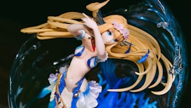 A detailed anime-style figure of a female character with long blonde hair and blue eyes, adorned with floral accessories and golden accents. Her attire is a mix of blue and white with seashell motifs, suggesting an oceanic or aquatic theme. She is surrounded by an abstract background resembling swirling water with intricate details.