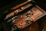 An ornately carved wooden jewelry box holds several pieces of elaborate jewelry. The jewelry features intricate designs with vibrant colors, including gold, red, and turquoise. The box and jewelry are illuminated by warm lighting, enhancing the rich details and colors.