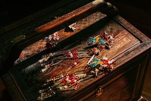 An ornately carved wooden jewelry box holds several pieces of elaborate jewelry. The jewelry features intricate designs with vibrant colors, including gold, red, and turquoise. The box and jewelry are illuminated by warm lighting, enhancing the rich details and colors.