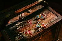 An ornately carved wooden jewelry box holds several pieces of elaborate jewelry. The jewelry features intricate designs with vibrant colors, including gold, red, and turquoise. The box and jewelry are illuminated by warm lighting, enhancing the rich details and colors.