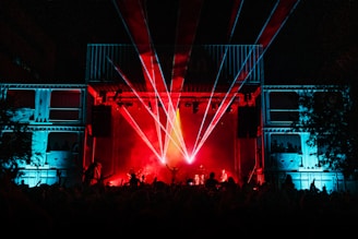 A lively concert scene with a large crowd in front of a stage, which is illuminated by vibrant red and blue lights. Laser beams are shooting out from the stage, adding to the dynamic and energetic atmosphere. The silhouettes of performers and audience members can be seen amidst the colorful lights.