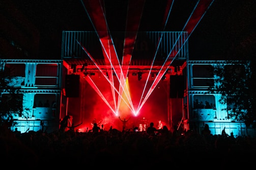 A lively concert scene with a large crowd in front of a stage, which is illuminated by vibrant red and blue lights. Laser beams are shooting out from the stage, adding to the dynamic and energetic atmosphere. The silhouettes of performers and audience members can be seen amidst the colorful lights.