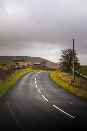 A winding asphalt road stretches through a rural landscape with grassy fields on either side. To the left, a stone barn is visible with a few black plastic-covered hay bales nearby. A bare tree stands on the right side next to a wooden pole. The sky is overcast with grey clouds, creating a moody atmosphere.
