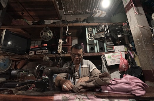 A man is focused on sewing using a vintage sewing machine in a cluttered workshop. The workspace is filled with various electronic equipment, reels, speakers, and a television. The walls are lined with items, including a calendar and a hanging plastic bag.