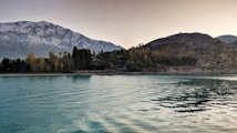 A serene lake with gentle ripples reflecting the soft light of the sky. Behind the lake, there are lush trees in autumn colors and a mountainous backdrop with snow-capped peaks. The sky is a gradient of warm hues, indicating either sunrise or sunset.