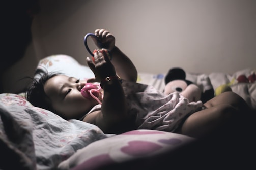 A baby lies on a bed, holding a teething ring with both hands. The baby has a pacifier in their mouth and wears a sleeveless romper with a bracelet on the right wrist. A plush toy, possibly Mickey Mouse, is seen in the background along with patterned sheets and pillows.