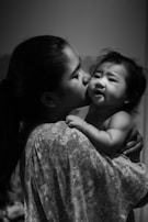 A woman gently holds and kisses a baby, creating a tender and intimate moment. The baby appears to be looking slightly away. The scene is captured in black and white, enhancing its emotional depth.