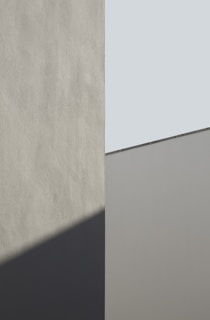 A minimalist architectural image featuring geometric shapes with smooth surfaces. The composition involves a vertical division, with one section casting a shadow on the adjoining wall. The colors involve muted tones and angles that create an interplay between light and shadow.