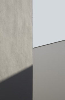 A minimalist architectural image featuring geometric shapes with smooth surfaces. The composition involves a vertical division, with one section casting a shadow on the adjoining wall. The colors involve muted tones and angles that create an interplay between light and shadow.
