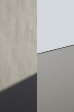 A minimalist architectural image featuring geometric shapes with smooth surfaces. The composition involves a vertical division, with one section casting a shadow on the adjoining wall. The colors involve muted tones and angles that create an interplay between light and shadow.